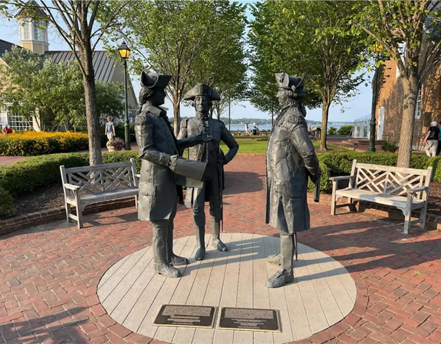 Three statues of 18th-century figures on a round platform in a brick-paved park area with trees and benches.